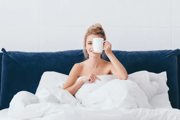 Belle femme émotionnelle avec tasse de café assis dans le lit le matin — Photo de stock