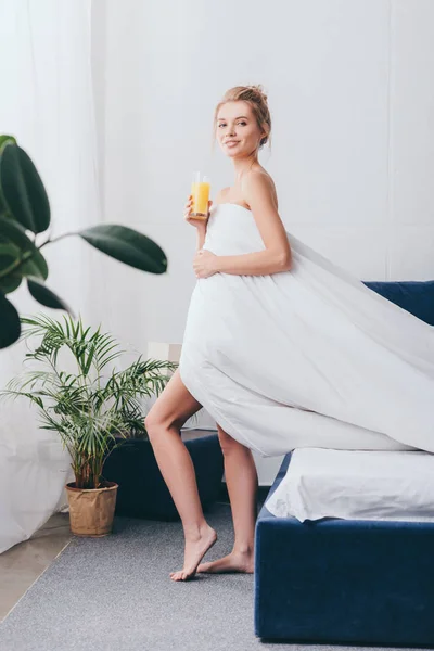 Bella giovane donna con bicchiere di succo in lenzuola bianche in camera da letto al mattino — Foto stock