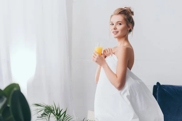 Donna sorridente con bicchiere di succo in lenzuola bianche in camera da letto al mattino — Foto stock