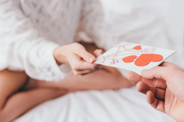 Vista cortada da mulher que toma cartão de saudação com corações para o dia dos namorados do homem na cama — Fotografia de Stock