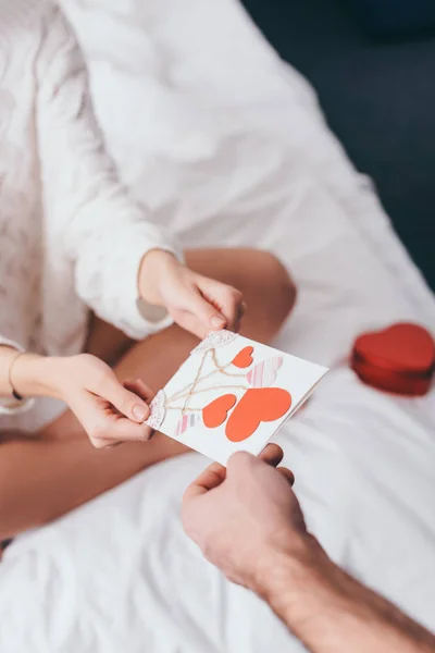 Обрезанный вид на пару с поздравительной открыткой и подарочной коробкой на день святого Валентина на кровати — стоковое фото