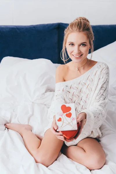 Attractive happy woman holding gift box and valentine card on bed — Stock Photo