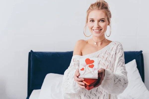 Belle femme souriante tenant boîte cadeau et carte de valentin sur le lit — Photo de stock