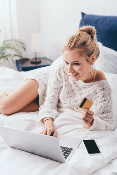 Belle femme heureuse shopping en ligne avec ordinateur portable et carte de crédit sur le lit le matin — Photo de stock