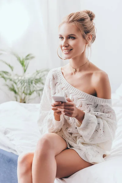 Hermosa mujer sonriente usando teléfono inteligente en la cama por la mañana - foto de stock