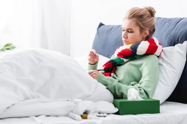 Donna malata in sciarpa con febbre e guardando il termometro a letto — Foto stock