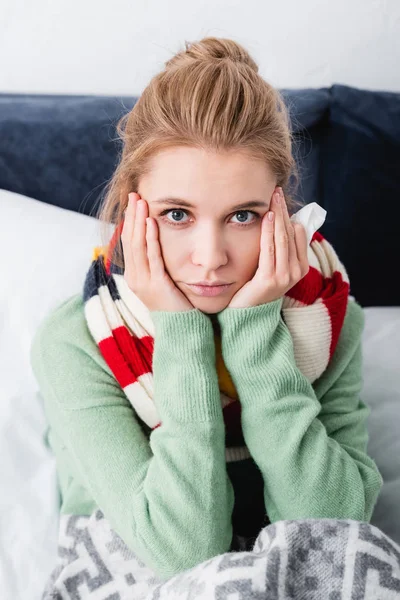 Sad ill woman in sweater and scarf holding napkin in bed — Stock Photo