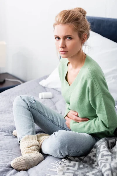 Sad woman having stomachache pain and sitting on bed with bottle of pills — Stock Photo