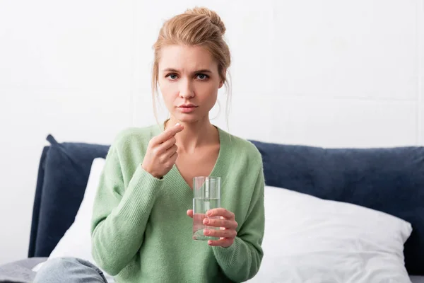 Mujer enferma triste tomando pastillas y sosteniendo un vaso de agua en el dormitorio - foto de stock