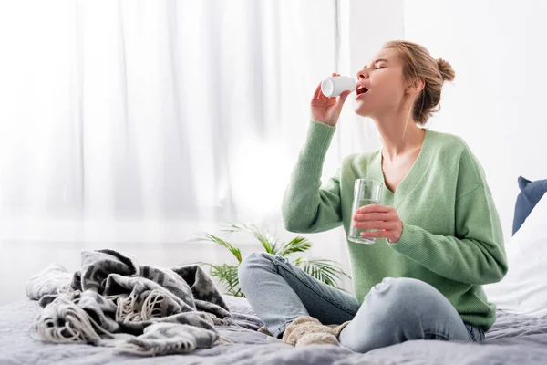 Donna malata che prende pillole dalla bottiglia e tiene un bicchiere d'acqua in camera da letto — Foto stock