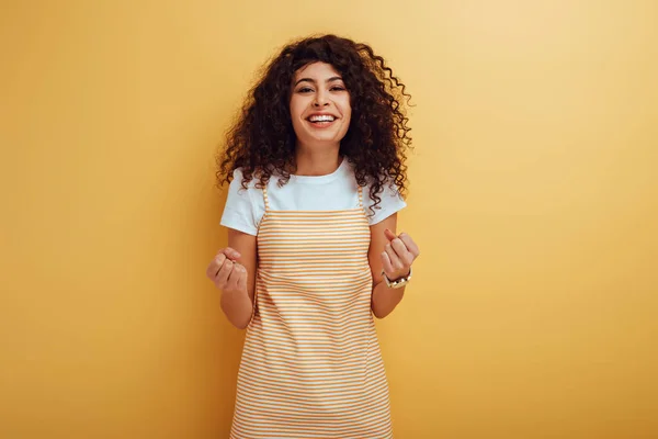 Happy mixed race girl showing winner gesture while looking at camera on yellow background — Stock Photo