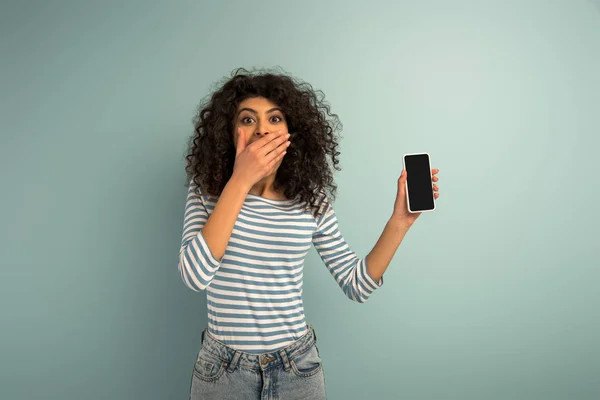 Impactada chica bi-racial que cubre la boca con la mano mientras muestra el teléfono inteligente con pantalla en blanco sobre fondo gris - foto de stock