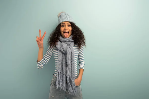 Alegre bi-racial chica en cálido sombrero y bufanda sobresaliendo lengua y mostrando el gesto de victoria sobre fondo gris - foto de stock