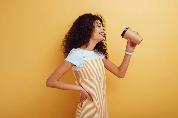 Alegre bi-racial chica bebiendo café para ir de pie con la mano en la cadera sobre fondo amarillo - foto de stock