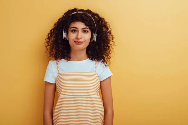 Bonita y sonriente chica bi-racial en auriculares inalámbricos mirando a la cámara en el fondo amarillo - foto de stock