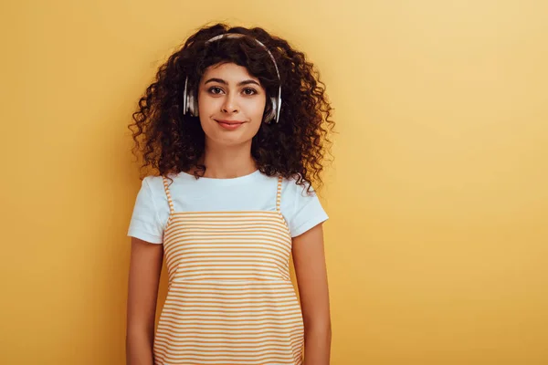 Jolie, souriante fille bi-raciale dans les écouteurs sans fil en regardant la caméra sur fond jaune — Photo de stock