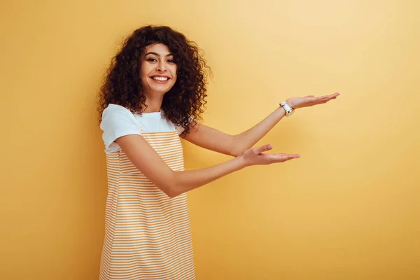 Felice ragazza bi-razziale che punta con le mani mentre sorride alla fotocamera su sfondo giallo — Foto stock