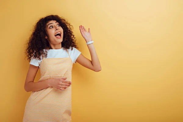 Felice bi-razziale ragazza agitando mano mentre guardando lontano su sfondo giallo — Foto stock