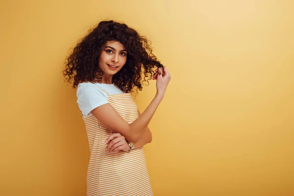 Paquera bi-racial menina tocando cabelo enquanto sorrindo para a câmera no fundo amarelo — Fotografia de Stock