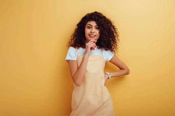Smiling bi-racial girl looking at camera while standing with hand on hip on yellow background — Stock Photo