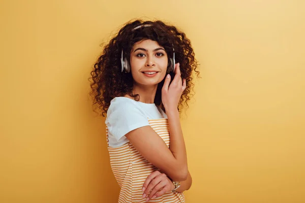 Smiling bi-racial girl in wireless headphones looking at camera on yellow background — Stock Photo