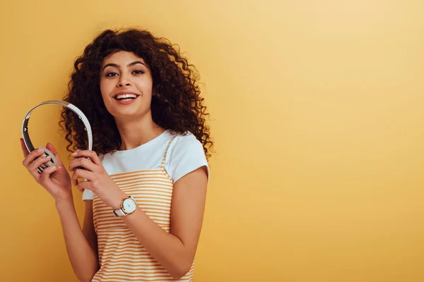 Feliz chica bi-racial sonriendo a la cámara mientras sostiene los auriculares inalámbricos en el fondo amarillo - foto de stock