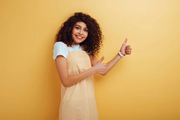 Alegre menina de raça mista mostrando polegares para cima enquanto olha para a câmera no fundo amarelo — Fotografia de Stock