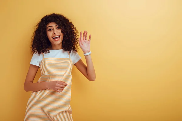 Happy mixed-race girl waving hand while looking at camera on yellow background — Stock Photo