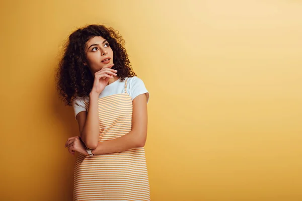 Nachdenkliche bi-rassische Frau schaut weg, während sie das Gesicht auf gelbem Hintergrund berührt — Stockfoto