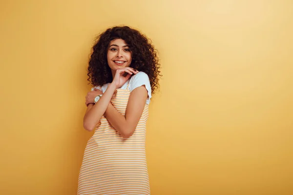 Cheerful bi-racial girl holding hand near face while looking at camera on yellow background — Stock Photo