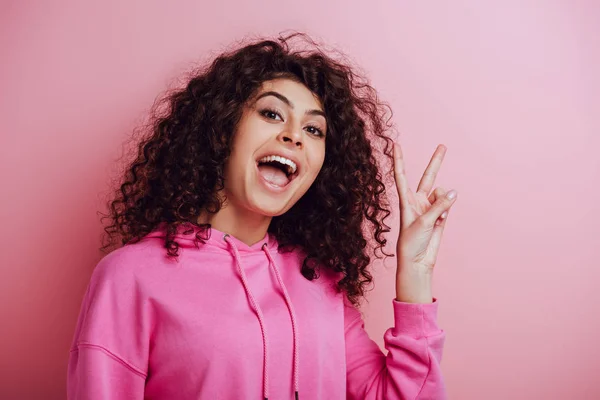 Excited bi-racial girl showing victory gesture while looking at camera on pink background — Stock Photo