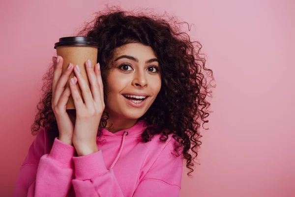 Alegre bi-racial chica sosteniendo café para ir mientras sonríe a la cámara en rosa fondo - foto de stock