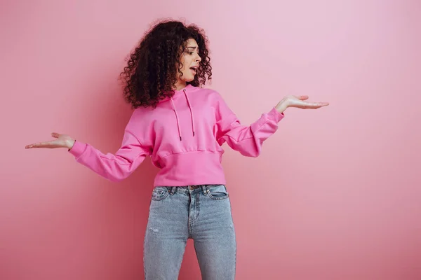 Displeased bi-racial girl standing with open arms on pink background — Stock Photo