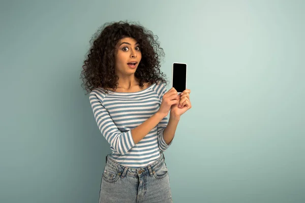 Sorprendida chica de raza mixta mostrando teléfono inteligente con pantalla en blanco sobre fondo gris - foto de stock