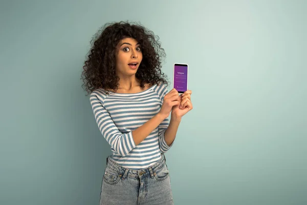 KYIV, UCRANIA - 26 de noviembre de 2019: chica de raza mixta sorprendida mostrando teléfono inteligente con aplicación Instagram sobre fondo gris - foto de stock