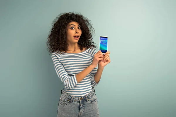 Sorprendida chica de raza mixta mostrando teléfono inteligente con aplicación de reserva sobre fondo gris — Stock Photo