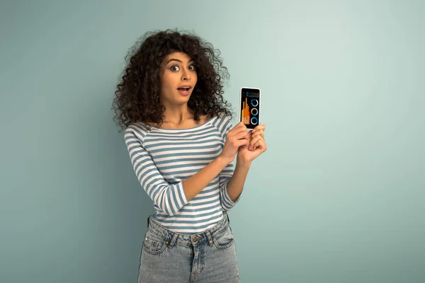 Sorprendida chica de raza mixta mostrando smartphone con gráficos y gráficos en pantalla sobre fondo gris - foto de stock