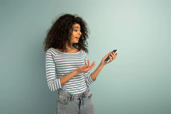 Displeased mixed race girl showing question gesture while looking at smartphone on grey background — Stock Photo