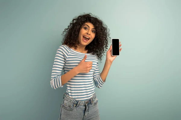 Glücklich gemischtes Rennmädchen zeigt Daumen nach oben, während es Smartphone mit leerem Bildschirm auf grauem Hintergrund hält — Stockfoto