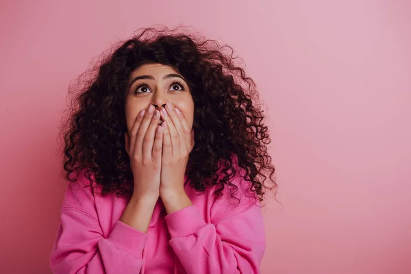 Choqué mixte race fille couvrant la bouche avec les mains tout en regardant vers le haut sur fond rose — Photo de stock