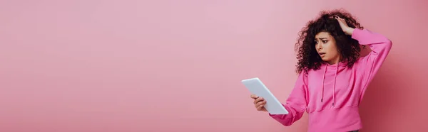 Panoramic shot of disappointed bi-racial girl touching hair while looking at digital tablet on pink background — Stock Photo