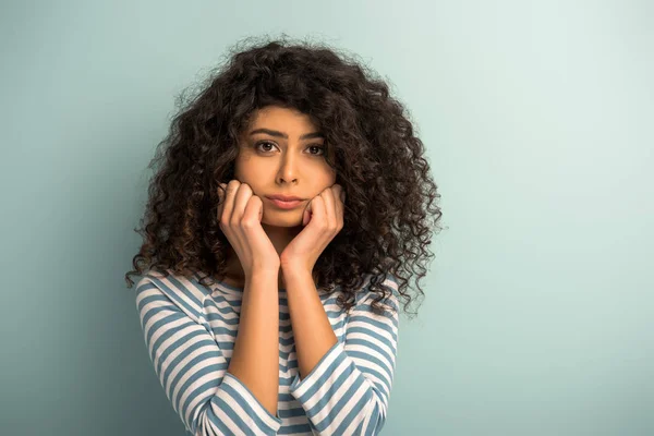 Disgustada chica de raza mixta mirando a la cámara mientras toma las manos cerca de la cara sobre fondo gris - foto de stock