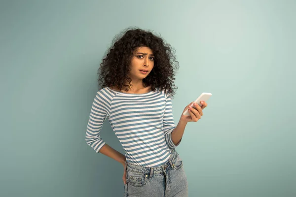 Displeased mixed race girl looking at camera while using smartphone on grey background — Stock Photo