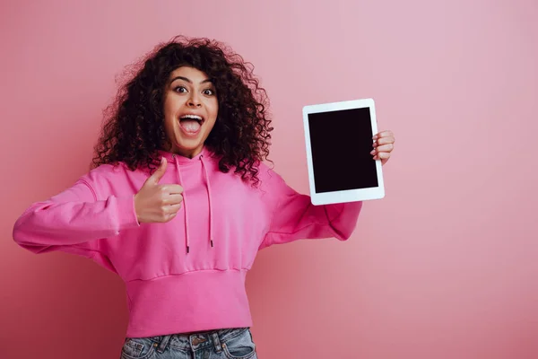 Alegre chica bi-racial sosteniendo tableta digital con pantalla en blanco y mostrando el pulgar hacia arriba sobre fondo rosa - foto de stock