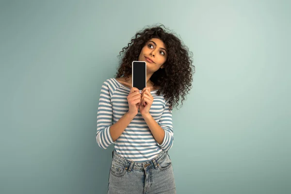 Pensativa chica de raza mixta mirando hacia otro lado mientras muestra el teléfono inteligente con pantalla en blanco sobre fondo gris - foto de stock