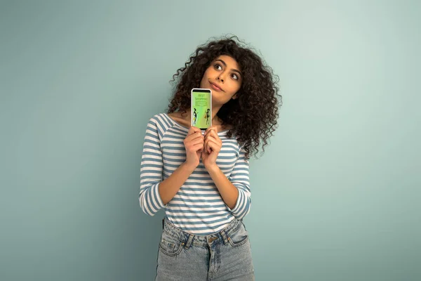 Menina de raça mista pensativa olhando para longe ao mostrar smartphone com o melhor aplicativo de compras no fundo cinza — Fotografia de Stock