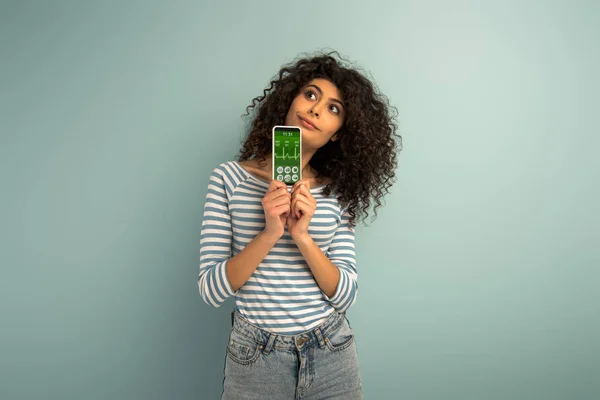 Reflexiva chica de raza mixta mirando hacia otro lado mientras muestra el teléfono inteligente con aplicación de rastreo de fitness en fondo gris - foto de stock