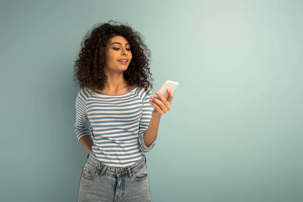 Улыбающаяся девушка смешанной расы болтает на смартфоне на сером фоне — стоковое фото