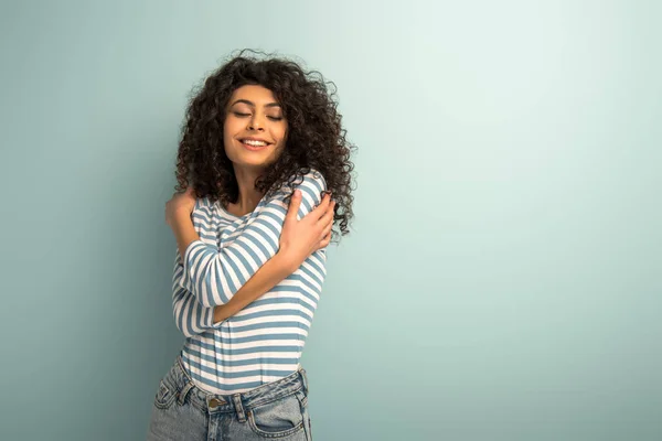 Happy mixed race girl hugging herself while standing with closed eyes on grey background — Stock Photo