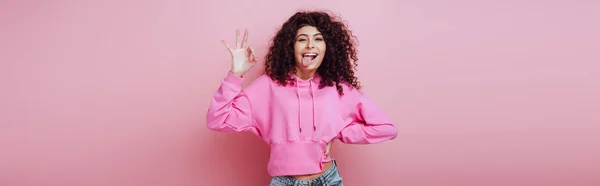 Plano panorámico de alegre chica bi-racial sobresaliendo lengua y mostrando gesto bien sobre fondo rosa - foto de stock